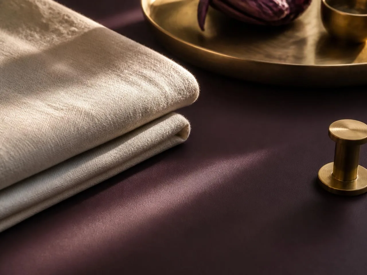 Folded cream linen towels and a brushed brass tray on a deep plum surface in warm, low afternoon light